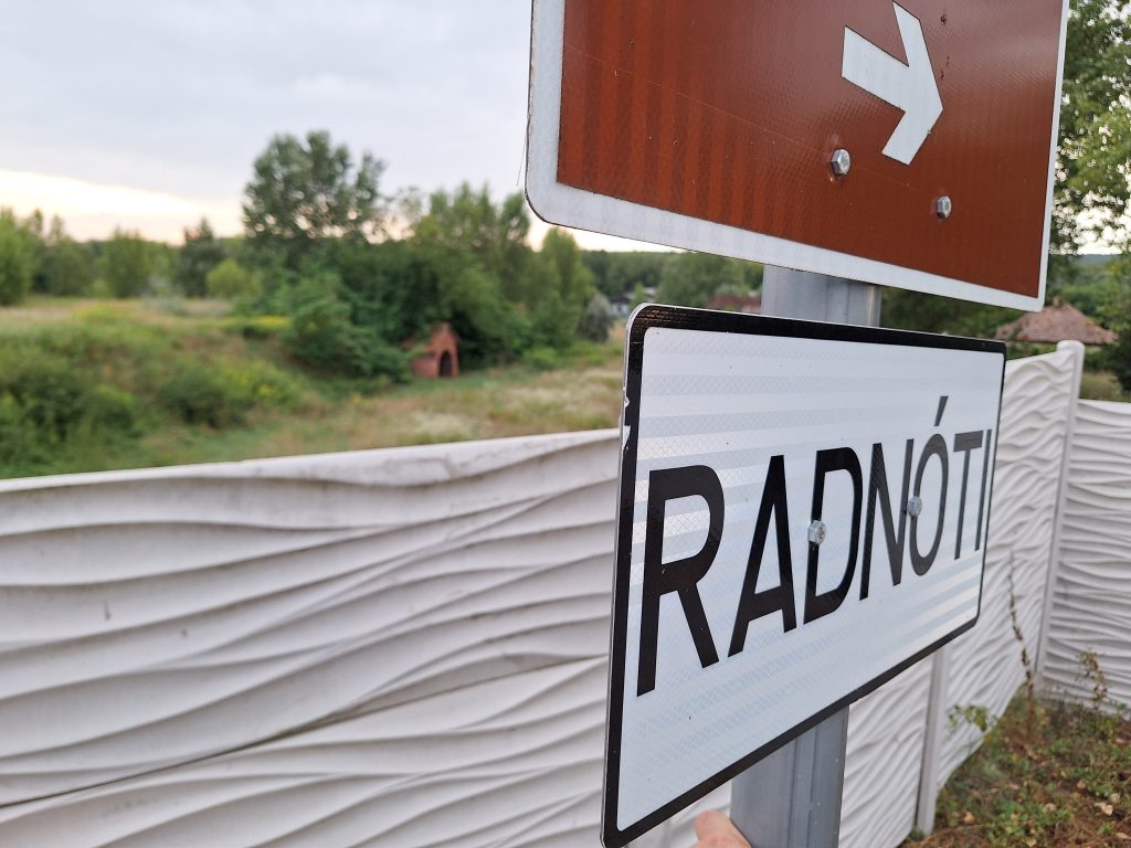 Az emlékművet jelző tábla, a kerítés fölött átkukkantva látszik a háttérben a meggyalázott emlékmű is